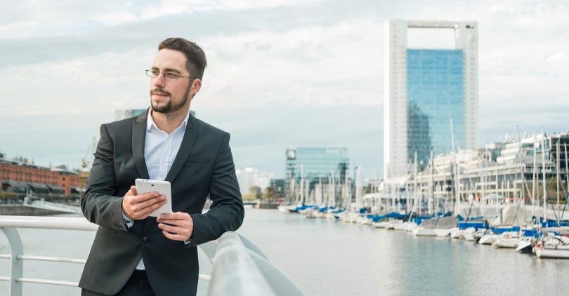 portrait-young-businessman-standing-near-harbor-holding-mobile-phone-hand-looking-away